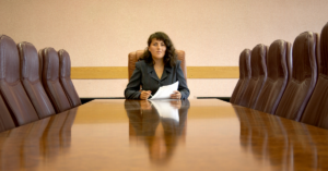 Woman sitting at the head of an empty board room table with papers in her hand.