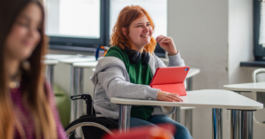 teenage wheelchair user with orange hair in a classroom with a tablet.